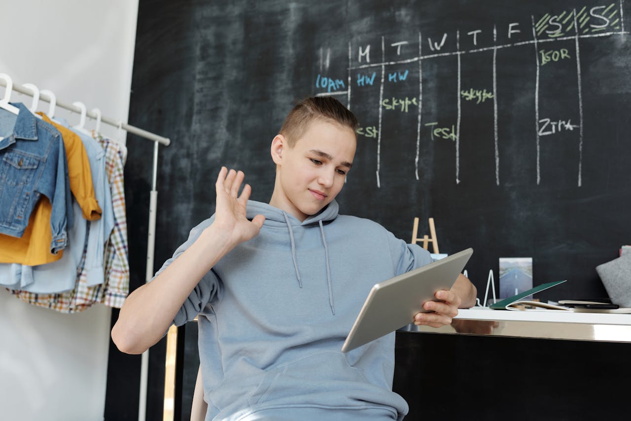 A teenager uses a tablet for online learning in a cozy home setting, waving at the screen.