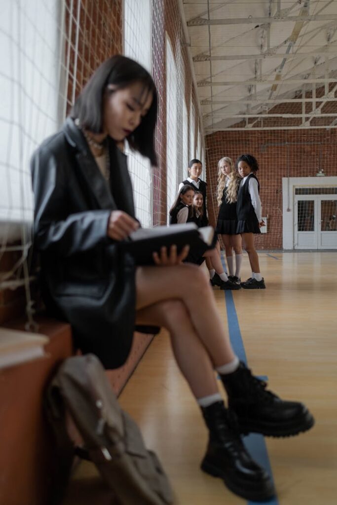 Teen girl reading alone in gymnasium while peers watch, highlighting themes of solitude and bullying.